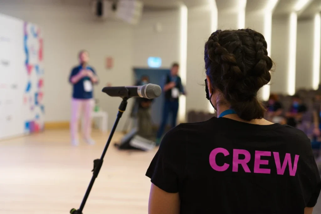 Me wearing a black shirt with “CREW” printed on it in purple stands near a microphone on a stand. The background is a blurred image of a speaker on stage and a few other people in a conference room setting.